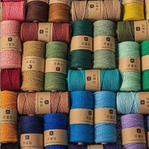 Könnte beinhalten: Eine Sammlung farbenfroher Garnrollen in verschiedenen Farbtönen, darunter Rosa, Braun, Grün, Blau und Gelb. Jede Rolle ist mit einem braunen Papierband umwickelt. Das Bild zeigt eine Vielzahl von Bastelbedarf.