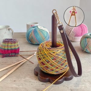 May include: A wooden yarn holder with a dark brown finish, holding a colorful ball of yarn. A brown leather wrist strap with a gold-colored clasp is attached. Knitting needles and other yarn balls are in the background, suggesting a crafting theme.