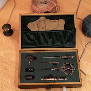May include: A wooden sewing kit with a green velvet interior. The kit contains ornate scissors, needles, a thimble, and other sewing tools. The kit is open, revealing the tools arranged neatly inside. The kit is on a wooden surface.