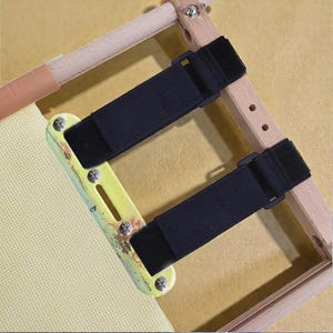 May include: A wooden embroidery hoop with black elastic straps and a yellow and green clip. The hoop is designed to hold fabric taut for cross-stitch or embroidery projects. The fabric is a light beige color.