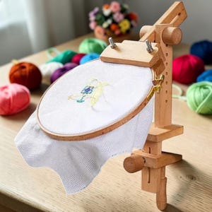 Può includere: Un supporto in legno per telaio da ricamo con tessuto bianco teso. Il supporto è in legno chiaro e ha elementi regolabili. Gomitoli di lana colorati sullo sfondo suggeriscono un tema artigianale.