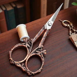 May include: Ornate copper-colored scissors with dragonfly and leaf designs, paired with a spool of beige thread. The scissors have sharp, silver blades and are set on a dark wooden surface, suggesting a craft or sewing theme.