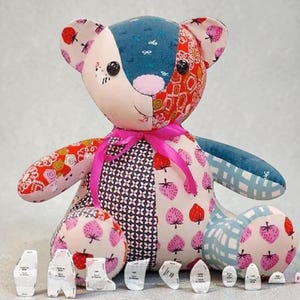 May include: A patchwork teddy bear constructed from various fabric patterns including strawberries, geometric shapes, and solid colors. The bear features black button eyes, a pink nose, and a bright pink ribbon tied around its neck. Small paper pattern pieces are arranged in a row in front of the bear.