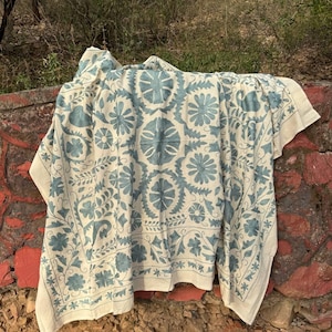 May include: A cream-coloured textile featuring intricate blue floral embroidery. The fabric displays a repeating pattern of large circular flower motifs surrounded by smaller leaves and vines, with a decorative border framing the entire design.