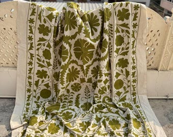 May include: A white textile with a green floral pattern. The design features large stylized flowers and leaves, with a border of smaller floral motifs. The fabric is draped over a surface, showcasing the intricate embroidery and color contrast.