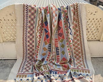 May include: Cream-colored textile with intricate embroidery in red, blue, green, and white. The design features geometric patterns and floral motifs. The textile is draped over a white structure, showcasing its detailed craftsmanship.