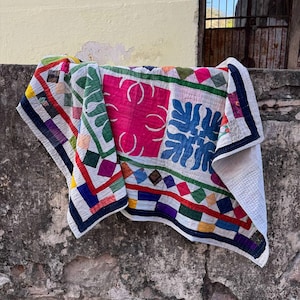 May include: A colorful patchwork quilt draped over a weathered wall. The quilt features a vibrant mix of pink, blue, green, and red geometric patterns. The edges are bordered with a dark blue fabric. The quilt is made of soft, textured fabric.