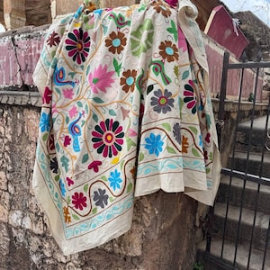 Può includere: Tessuto beige con vivaci ricami floreali in rosa, blu, verde e marrone. Il design ricamato include fiori, foglie e uccelli. Il tessuto è drappeggiato su una struttura in pietra, mettendo in risalto l'intricata lavorazione artigianale.