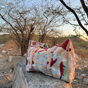 Puede incluir: Un bolso tote grande y colorido con un diseño de patchwork, con patrones rojos, naranjas y azules. La bolsa está sobre una gran roca al aire libre, con árboles y un paisaje natural al fondo.