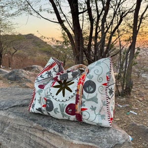 Puede incluir: Un bolso tote blanco con un estampado floral en rojo, verde y marrón, apoyado sobre una gran roca. El bolso tiene forma rectangular con un borde estampado y un asa. El fondo presenta árboles y una puesta de sol.