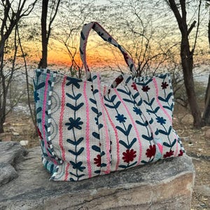 Puede incluir: Un bolso tote grande con un estampado floral en tonos rosa, azul y rojo sobre un fondo crema. El bolso tiene un asa a juego y está colocado sobre una gran roca. El fondo presenta árboles y una puesta de sol.