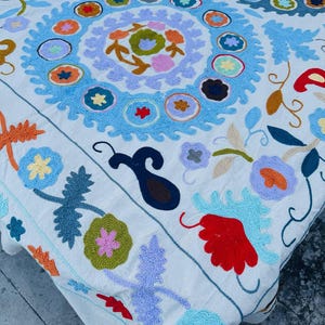 Può includere: Tessuto bianco con ricami intricati multicolori. Il design presenta motivi floreali e geometrici, inclusi motivi circolari e fiori stilizzati nelle tonalità di blu, arancione, rosso e verde. Il tessuto sembra essere un oggetto decorativo.