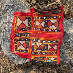 Könnte beinhalten: Ein leuchtend rotes Textil mit aufwendigen Stickereien und Spiegelakzenten. Das Design zeigt geometrische Muster in Gelb, Weiß und Rot mit einem schwarzen Rand. Dieses handgefertigte Stück zeigt traditionelle Handwerkskunst und kulturelle Kunst.