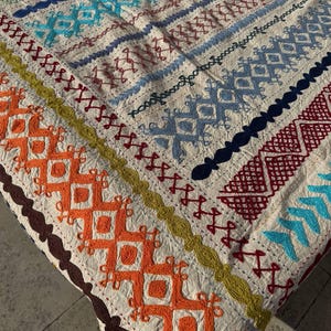 Può includere: Tessuto color crema con intricati motivi ricamati in vari colori. Il design include forme geometriche, linee e dettagli a punto croce nelle tonalità dell'arancione, blu, verde e bordeaux. I bordi sono rifiniti con un bordo marrone.