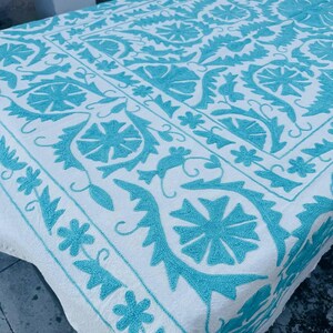 May include: White textile with detailed turquoise embroidery. The design features floral and geometric patterns, creating an ornate appearance. The embroidery is evenly spaced across the fabric, with a border of repeating shapes.