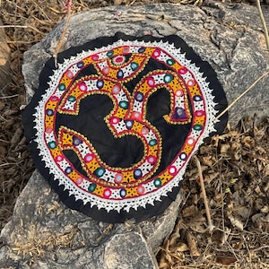 Può includere: Tessuto rotondo nero con un simbolo Om centrale ricamato in giallo, rosso e bianco. Il simbolo è circondato da un bordo di perline colorate e un bordo di pizzo bianco. Il tessuto è posto su una roccia grigia.