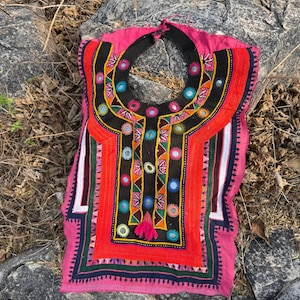 Könnte beinhalten: Eine rosa, rote und schwarze bestickte Tunika mit rundem Ausschnitt. Die Tunika hat farbenfrohe geometrische Muster, darunter Kreise und Dreiecke, und eine kleine Quaste. Das Kleidungsstück wird auf einer natürlichen Oberfläche im Freien präsentiert.