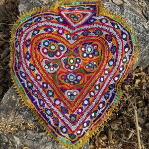 Puede incluir: Textil bordado en forma de corazón con un diseño vibrante. La pieza presenta una base morada, adornada con costuras rojas, blancas y doradas, y pequeños acentos de espejo. Los bordes están ribeteados con flecos dorados.