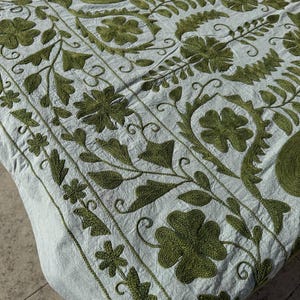 Könnte beinhalten: Hellblaues Textil mit detaillierter olivgrüner Stickerei. Das Design zeigt florale und Paisley-Muster mit strukturierten Nähten. Dieses Textil ist wahrscheinlich ein dekorativer Artikel, wie ein Überwurf oder eine Tischdecke, der einen Hauch von Eleganz verleiht.