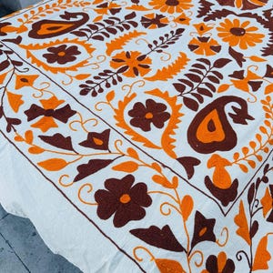 Può includere: Tessuto bianco con ricami intricati nei toni del marrone e dell'arancione. Il design presenta motivi floreali e paisley, creando una superficie decorativa. Il ricamo è uniformemente distanziato sul tessuto, mostrando un artigianato tradizionale.