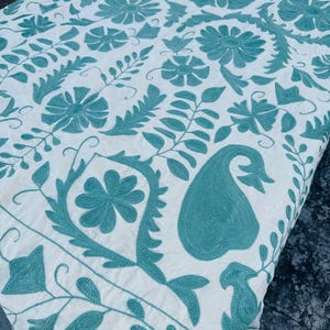 Peut inclure: Textile blanc avec broderie florale turquoise. Le motif présente de grandes fleurs, des feuilles et d'autres éléments botaniques. La broderie est détaillée et couvre toute la surface du tissu. Le textile semble être une courtepointe ou un type de literie similaire.
