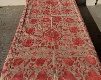 May include: A beige textile with intricate red floral embroidery. The design features large flowers, paisley-like shapes, and scrolling patterns. The textile is likely a decorative bedspread or wall hanging.