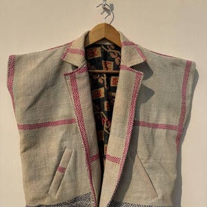 Könnte beinhalten: Eine ärmellose, beige Jacke mit gemustertem Innenfutter und rosa Besatz. Die Jacke hat ein maßgeschneidertes Design mit einem Reverskragen und zwei Vordertaschen. Die Kanten sind mit einem dunkelblauen und weißen geometrischen Muster akzentuiert.