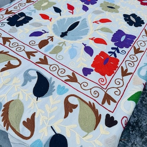 Könnte beinhalten: Weißes Textil mit aufwendiger Blumenstickerei. Die Stickerei umfasst rote, violette, blaue und grüne Blumen sowie Paisley- und Blattmotive. Der Rand hat ein dekoratives, wirbelndes Muster.