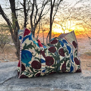 Puede incluir: Un cojín decorativo con un estampado floral en tonos azules, rojos y verdes sobre un fondo crema. El cojín está colocado sobre una superficie de piedra, con una puesta de sol al fondo. El cojín tiene un borde a rayas.