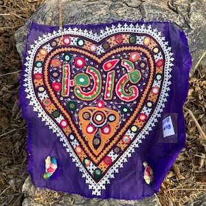 Può includere: Un tessuto viola con un disegno a forma di cuore. Il cuore è riempito di ricami colorati, specchi e il numero "1516" in verde, rosso e arancione. Un bordo bianco ricamato circonda il cuore. Piccoli abbellimenti colorati sono sugli angoli.