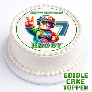 Può includere: Una torta rotonda bianca con un bordo verde e un'immagine colorata a tema Roblox. L'immagine presenta un personaggio dei cartoni animati che tiene in mano un controller di gioco, con il nome "BRODY" e il numero "7". La torta è decorata con glassa bianca e le parole "HAPPY BIRTHDAY".