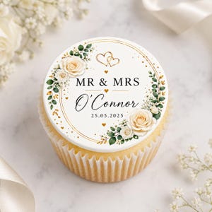 Op de afbeelding: Een cupcake met een witte frosting schijf met de tekst "MR & MRS O'Connor" en de datum "25.05.2025". De schijf is versierd met gouden accenten, rozen en groen. De cupcake zit in een lichtbruine papieren cup.