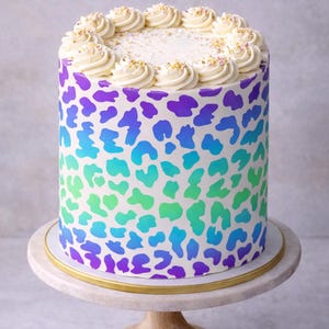 Pode incluir: Um bolo redondo com uma base de glacê branco, decorado com um padrão de leopardo colorido em tons de roxo, azul e verde. O topo é adornado com redemoinhos de glacê branco e granulado. O bolo está sobre um suporte de bolo de madeira.