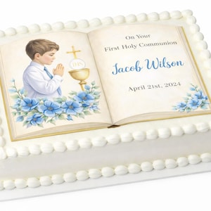 Può includere: Una torta bianca decorata con un'immagine stampata di un libro aperto, un ragazzo che prega e fiori blu. Il libro recita "On Your First Holy Communion" e il nome "Jacob Wilson" con la data "April 21st, 2024".