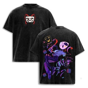 May include: A black acid-wash t-shirt featuring a small white and red theatrical mask graphic on the front chest. The back displays a large, colorful illustration of a character with a mask, surrounded by floating ribbons and small circular objects.