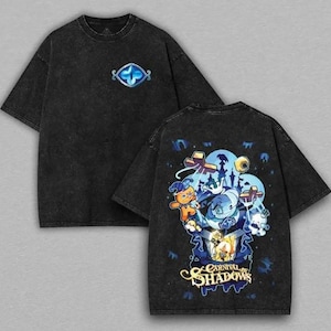 May include: A black acid-wash t-shirt featuring a small blue eye graphic on the front chest. The back displays a large, colorful illustration of various cartoon characters and the text "CARNIVAL OF SHADOWS" in a stylized, golden-yellow font.