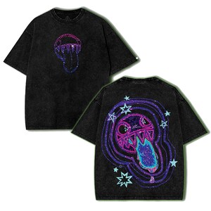 May include: A black acid-wash t-shirt featuring a small graphic on the front and a large graphic on the back. Both graphics depict a stylized creature with sharp teeth and a long tongue, rendered in vibrant purple, pink, and blue glittery tones.