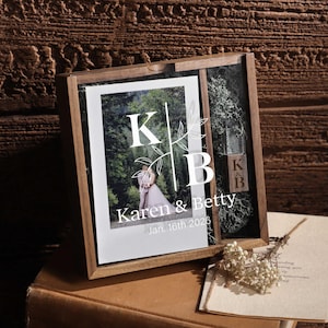 May include: A wooden shadow box with a wedding photo and decorative elements. The photo features a couple in a forest setting, with the names "Karen & Betty" and the date "Jan. 16th 2026" printed below. The box also includes initials "K & B" on a small block.