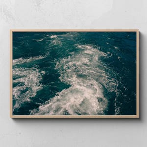 Puede incluir: Una impresión enmarcada que presenta una vista de cerca del agua del océano en movimiento. El agua es de un color verde azulado profundo, con espuma blanca y patrones arremolinados. El marco de madera tiene un acabado natural.