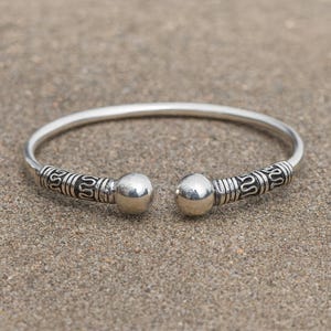 May include: A silver cuff bracelet with etched patterns near the ends. The bracelet has a smooth, rounded band and two spherical accents. The bracelet is displayed on a textured, light brown surface.