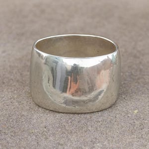May include: A wide, polished silver ring with a smooth, rounded design. The ring has a thick band and a simple, minimalist style. The reflective surface of the ring catches the light, creating highlights and shadows. This piece of jewelry is suitable for everyday wear.