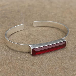 May include: A silver cuff bracelet featuring a rectangular, deep red gemstone. The bracelet has a simple, modern design with a polished finish. The open design allows for easy wear.