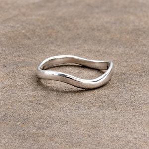 Może przedstawiać: Srebrny pierścionek z falistym wzorem. Pierścionek ma gładkie, polerowane wykończenie i płynny, organiczny kształt. Pierścionek jest prezentowany na neutralnej, teksturowanej powierzchni.