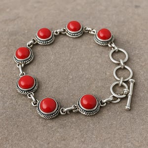 May include: A silver bracelet featuring eight round, bright red stones. Each stone is set in a detailed, beaded silver bezel. The bracelet has a toggle clasp closure. The jewelry is displayed on a neutral surface.