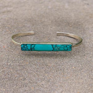 May include: A silver cuff bracelet with a rectangular turquoise stone. The turquoise stone is a vibrant blue-green colour with dark veining. The bracelet has a simple, minimalist design.