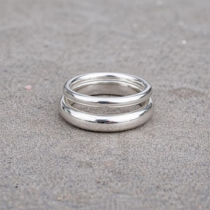 May include: A pair of silver rings, one stacked on top of the other, creating a double-banded effect. The rings are smooth and shiny, reflecting the light. The rings are set against a gray, textured background.