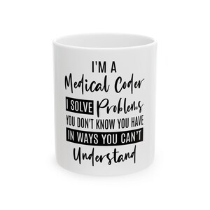 Op de afbeelding: Witte keramische mok met de tekst "I'M A Medical Coder I SOLVE Problems YOU DON'T KNOW YOU HAVE IN WAYS YOU CAN'T Understand" in zwart schrift. De mok is cilindrisch en heeft een eenvoudig, strak ontwerp.