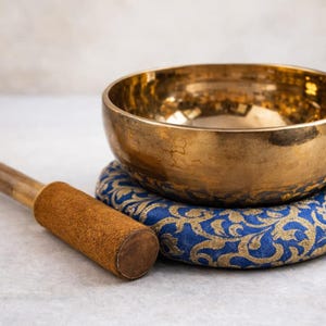 Peut inclure: Un bol chantant doré posé sur un coussin bleu et or à motifs, avec un maillet en bois à côté. Le bol a une surface lisse et réfléchissante. Le maillet a une poignée en daim marron. Cet article est utilisé pour la méditation et la guérison sonore.