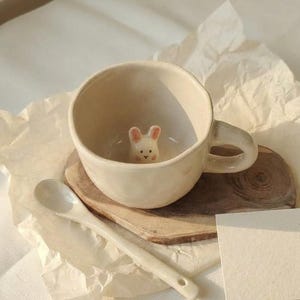 Op de afbeelding: Crèmekleurige keramische mok met een konijnenbeeldje erin, een bijpassende lepel en een kleine houten onderzetter. De mok heeft een oor en staat op een stuk hout. Een blanco kaart en gekreukeld papier zijn ook aanwezig.