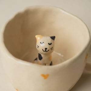 Może przedstawiać: Kremowy ceramiczny kubek z małą, ręcznie malowaną figurką kota w środku. Kot ma czarne, żółte i białe oznaczenia. Kubek ma na zewnątrz wzór serca.